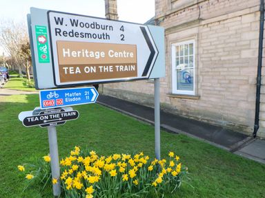 Road signs with directions to W Woodburn Redesmouth Heritage Centre and Tea on the Train near a building with daffodils in the grass at Rose Hip Cottage in Bellingham