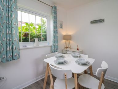 A dining area with a white table set for four and light blue curtains with seagull prints at Sea Rose in Aberporth