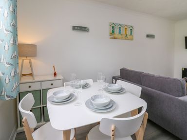 A dining table set with plates bowls and glasses next to a cabinet with a lamp and a curtain with bird pattern at Sea Rose in Aberporth