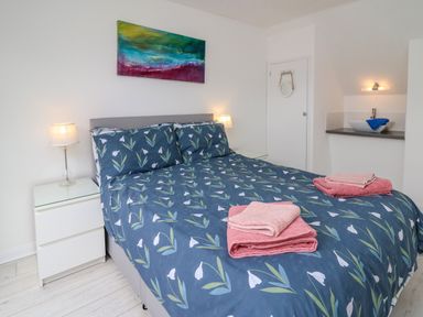 A bedroom with a double bed with floral bedding two pink towel sets on the bed bedside tables with lamps and a washbasin with a mirror on the wall at Sea Rose Aberporth