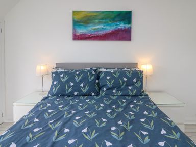 A bedroom with a double bed with floral bedding two bedside tables with lamps and a colorful abstract painting on the wall at Sea Rose in Aberporth