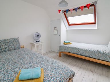 A bedroom with two single beds with patterned bedding a white nightstand with a fan and a window with triangular bunting at Sea Rose in Aberporth