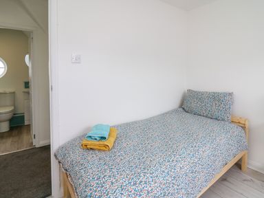 A bedroom with a single bed covered with patterned bedding and folded towels on the bed next to a doorway showing a bathroom with a toilet and sink at Sea Rose in Aberporth