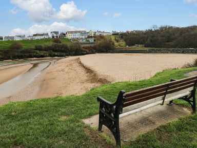 A wooden bench on grass overlooking a sandy beach and houses on a hill in the background at Sea Rose Aberporth