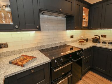 A kitchen with black cabinets a marble countertop an electric stove a cookbook a cake under a glass dome a kettle and three mugs at Manor Farm House in Bere Regis