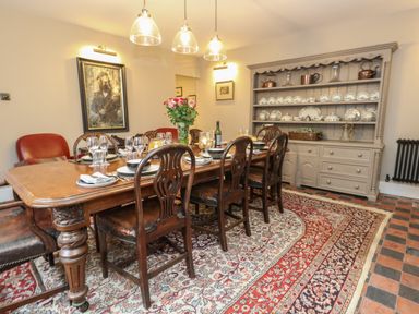 A dining room with a long wooden table set with dishes and glasses surrounded by wooden chairs and a large cabinet with plates in Bere Regis