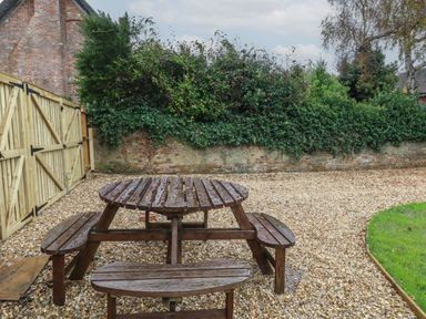 A wooden picnic table on gravel with a wooden fence and brick wall covered in greenery at Manor Farm House in Bere Regis