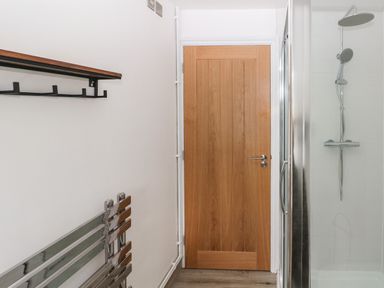 A bathroom with a wooden door a tiled shower area with a silver showerhead a metallic heated towel rail and a wall-mounted shelf with hooks at Folly Cottage in Llanteg near Saundersfoot