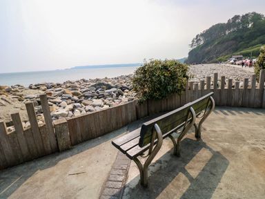 A wooden bench facing a pebble beach and sea with a wooden fence and rocky hill in the background at Folly Cottage in Llanteg near Saundersfoot