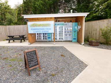 An outdoor vending area with machines and a picnic bench surrounded by a wooden fence at Pembrokeshire Fresh Llanteg Vending