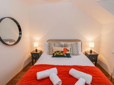 A bedroom with a bed with pillows and a red blanket with towels on the bed and two bedside tables with lamps and a round mirror on the wall at Folly Cottage in Llanteg near Saundersfoot