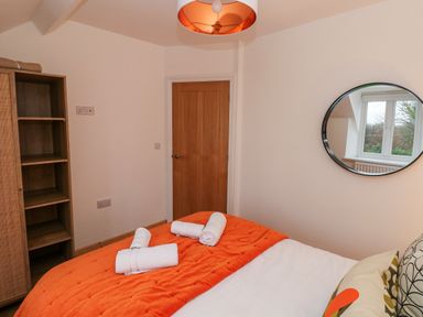 A bedroom with an orange bedspread and rolled towels on the bed a wooden door a wooden shelf unit and a round mirror on the wall reflecting a window at Folly Cottage in Llanteg near Saundersfoot