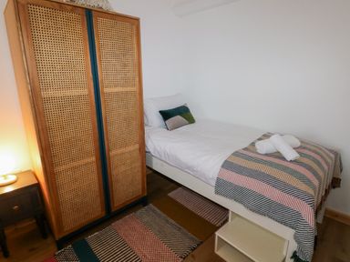 A bedroom with a single bed covered by a striped blanket and two rolled towels on top a wooden wardrobe a bedside table with a lamp and a patterned rug on the floor at Folly Cottage in Llanteg near Saundersfoot