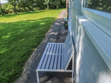 A metal bench beside the wall of a house on a paved pathway with grass and trees in the background at 61 Norton Park in Dartmouth