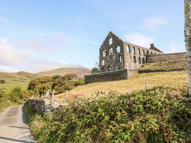 A rural road next to a stone wall with ruins of a building on a grassy hill and hills in the background at Ereiniog 2 in Tremadog