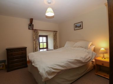 A bedroom with a double bed between a wooden chest of drawers and a wooden nightstand with a lamp and a small window with curtains at Ereiniog 2 in Tremadog