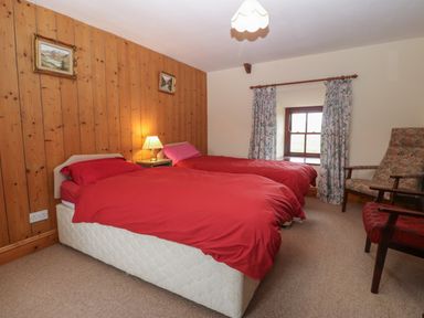 A bedroom with two single beds with red bedding a wood paneled wall paintings a bedside table with lamp a window with floral curtains and two armchairs at Ereiniog 2 in Tremadog