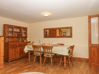 A dining room with a wooden table covered by a polka dot tablecloth surrounded by six wooden chairs with a wooden cabinet displaying dishes and a mirror on the wall at Peace Cottage Queens Nympton near South Molton