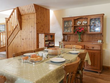 A dining room with a polka dot tablecloth on a table set with plates cups and croissants and a wooden cabinet with dishes and flowers at Peace Cottage in Queens Nympton near South Molton