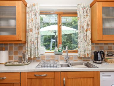 A kitchen sink with dish rack and cleaning supplies under a window with curtains and wooden cabinets at Peace Cottage in Queens Nympton near South Molton