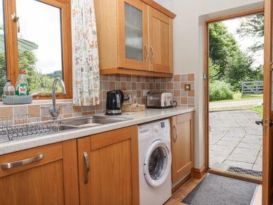 A kitchen with wooden cabinets a washing machine sink with faucet a kettle toaster and an open door showing an outside path at Peace Cottage Queens Nympton near South Molton