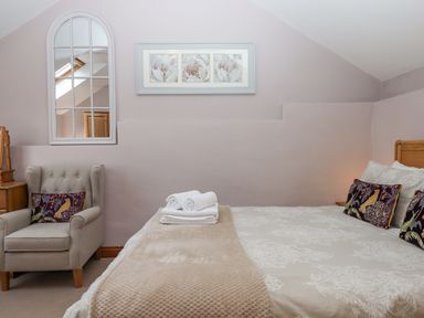 A bedroom with a bed with folded towels and cushions a grey armchair with a cushion a wooden bedside table and a wall mirror at Peace Cottage in Queens Nympton near South Molton