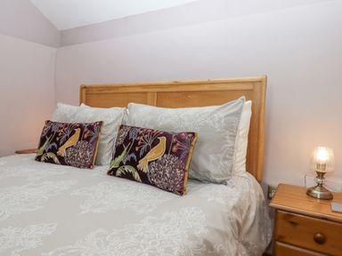 A bedroom with a wooden bed frame a patterned bedspread two bird design pillows and a nightstand with a lamp at Peace Cottage Queens Nympton near South Molton