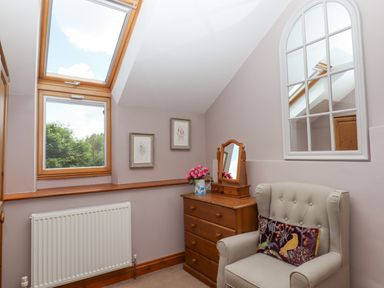A corner of a room with a beige armchair and a wooden chest of drawers with a mirror and flowers on top at Peace Cottage in Queens Nympton near South Molton