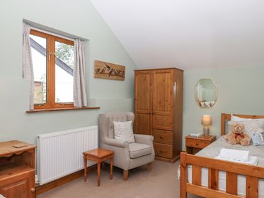 A bedroom with wooden furniture including a bed wardrobe and side tables a grey armchair and a window with curtains at Peace Cottage Queens Nympton near South Molton