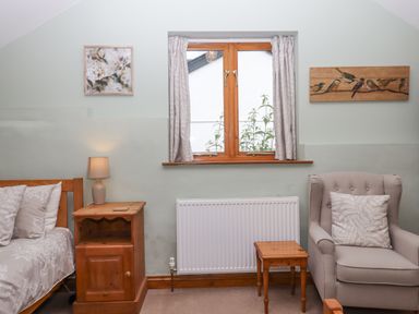 A bedroom with a wooden bed side table with lamp a beige armchair wooden side table and a window with curtains at Peace Cottage Queens Nympton near South Molton