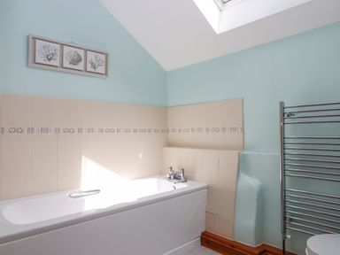 A bathroom with a white bathtub tiled walls a skylight a towel radiator and framed artwork at Peace Cottage Queens Nympton near South Molton