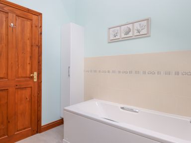 A bathroom with a wooden door a white storage cabinet a white bathtub and three framed pictures above the bathtub at Peace Cottage in Queens Nympton near South Molton