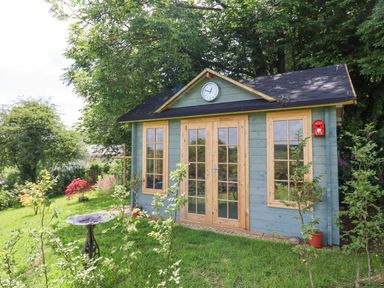 A small blue wooden garden shed with glass paneled doors and windows surrounded by grass and trees at Peace Cottage Queens Nympton near South Molton