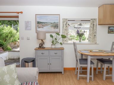 A dining area with a round wooden table and chairs a sideboard with a plant and lamp and a window with floral curtains at Dove Cottage Queens Nympton near South Molton