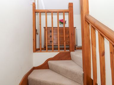 A staircase with beige carpet and wooden railing leading to a landing with wooden chest of drawers and pink flowers in a vase at Dove Cottage Queens Nympton near South Molton