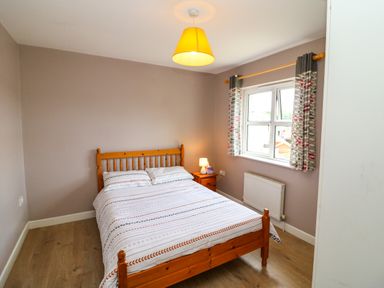 A bedroom with a wooden bed and bedside table with lamp curtains on the window at 23 Cois Caol in Skibbereen County Cork