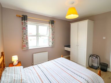 A bedroom with a bed a bedside table with a lamp curtains on the window a white wardrobe and laundry and rubbish bins at 23 Cois Caol in Skibbereen County Cork