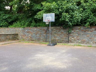 An outdoor basketball hoop on a paved surface with a stone wall and trees in the background at 23 Cois Caol in Skibbereen County Cork