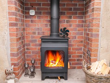 A wood burning stove with fire inside surrounded by brick walls and a basket of firewood at Woodcutters Cottage in Park Village near Haltwhistle