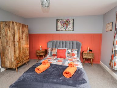 A bedroom with a bed a grey headboard orange and grey bedding two wooden bedside tables with lamps a wooden wardrobe and a painting on the wall at Woodcutters Cottage Park Village near Haltwhistle