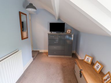 A small room with a grey cabinet holding a television and picture frame a wooden chest with three picture frames a wall mirror a radiator and a pendant light at Woodcutters Cottage in Park Village near Haltwhistle