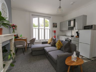 A living area with a gray sofa, two chairs, a small table with tissues, a kitchen with white cabinets, a refrigerator, microwave, and a window with white blinds at Flat 2 - Studland in Bournemouth