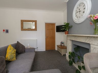 A living room with a gray sofa yellow and gray cushions a wooden door a wall clock plants and a marble fireplace at Flat 2 - Studland in Bournemouth