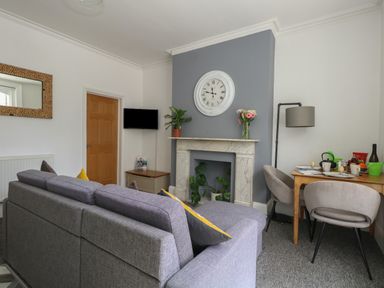 A living room with a gray sofa a wooden door a wall clock on a gray accent wall above a fireplace and a dining table with chairs and a floor lamp at Flat 2 - Studland in Bournemouth