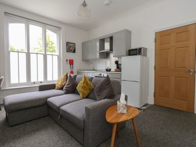 A living area with a gray sofa and a small wooden table in front of a kitchen with gray cabinets and a white refrigerator at Flat 2 - Studland in Bournemouth
