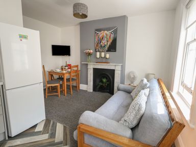 A living area with a gray sofa, a wooden dining table with chairs, a fireplace with candles, a colorful cow painting, a white refrigerator, and a window at Flat 4 - Hengistdury in Bournemouth
