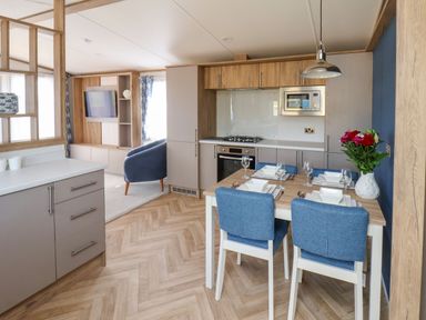 A kitchen and dining area with a wooden floor a table set for four with blue chairs and a living area with a blue armchair and a wall-mounted tv at Juniper Lodge in Whitby