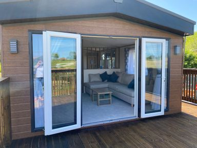A wooden lodge with white framed glass doors opening to a living room with a corner sofa and coffee table at Willow Lodge in Whitby