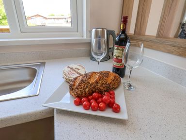 A kitchen countertop with a loaf of bread and cherry tomatoes on a plate with a bottle of wine two empty wine glasses a container of cheese and a kettle at Magnolia Lodge in Whitby