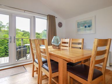 A dining room with a wooden table and six chairs next to glass doors showing a balcony with outdoor furniture at 3 Lake View in Lanreath